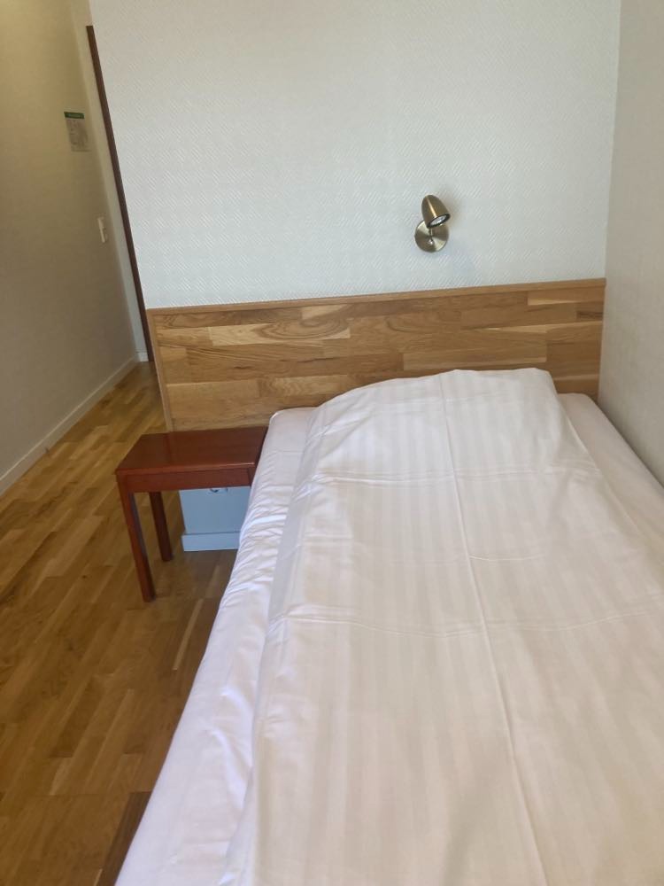 Pokój hotelowy z pojedynczym łóżkiem, drewnianym zagłówkiem i stolikiem nocnym, oświetlony kinkietem