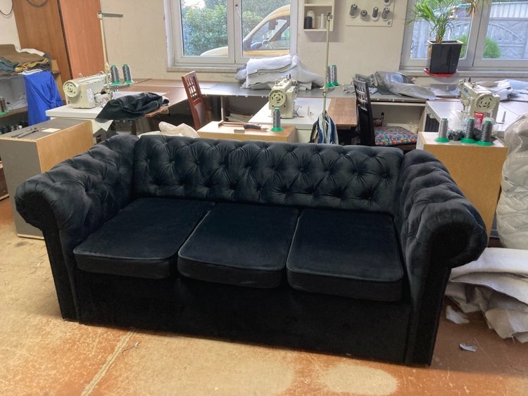 Czarna, welurowa sofa typu Chesterfield w warsztacie, widoczne maszyny do szycia i materiały tapicerskie w tle.