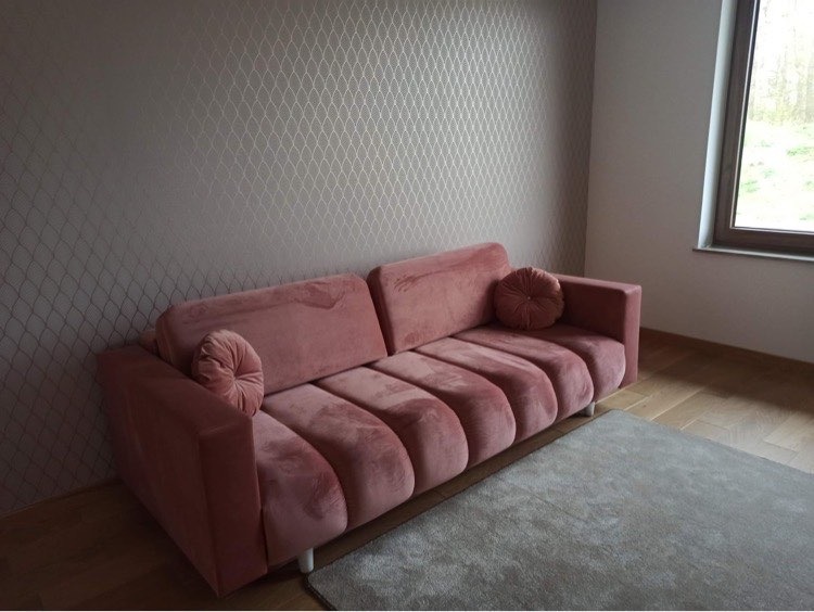 Różowa sofa z weluru z okrągłymi poduszkami dekoracyjnymi na tle ściany w geometryczny wzór i jasnego dywanu.