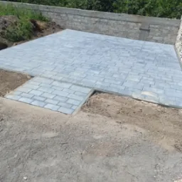 Ułożona, szara kostka brukowa na podjeździe, widoczne niedokończone krawędzie i ziemia, tło stanowi mur oporowy i zieleń.