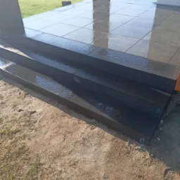 Nowo wykonane, czarne schody tarasowe z błyszczących, prostokątnych elementów, prowadzące na taras z szarych, kwadratowych płyt, widoczne ślady wilgoci, w tle trawnik i zwinięty wąż ogrodowy.