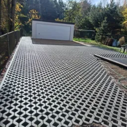 Ułożona nawierzchnia z ażurowych płyt betonowych prowadząca do białego garażu, widok z góry, z boku metalowe ogrodzenie i drzewa w tle.