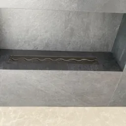 Wnęka w ścianie wyłożona szarymi płytkami imitującymi kamień, z wbudowanym biokominkiem z metalową listwą palnika o falistym wzorze.