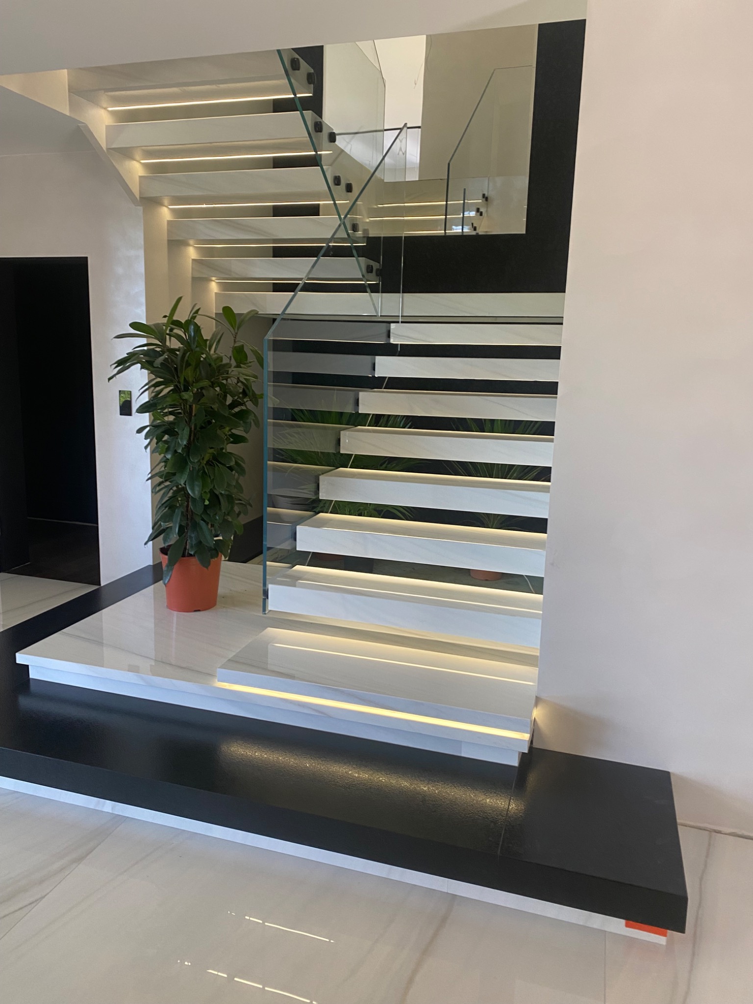 Nowoczesne schody z białego marmuru z podświetleniem LED, szklaną balustradą i rośliną doniczkową w pomarańczowej donicy. Widoczny fragment czarnej ściany i podłogi z białego marmuru.