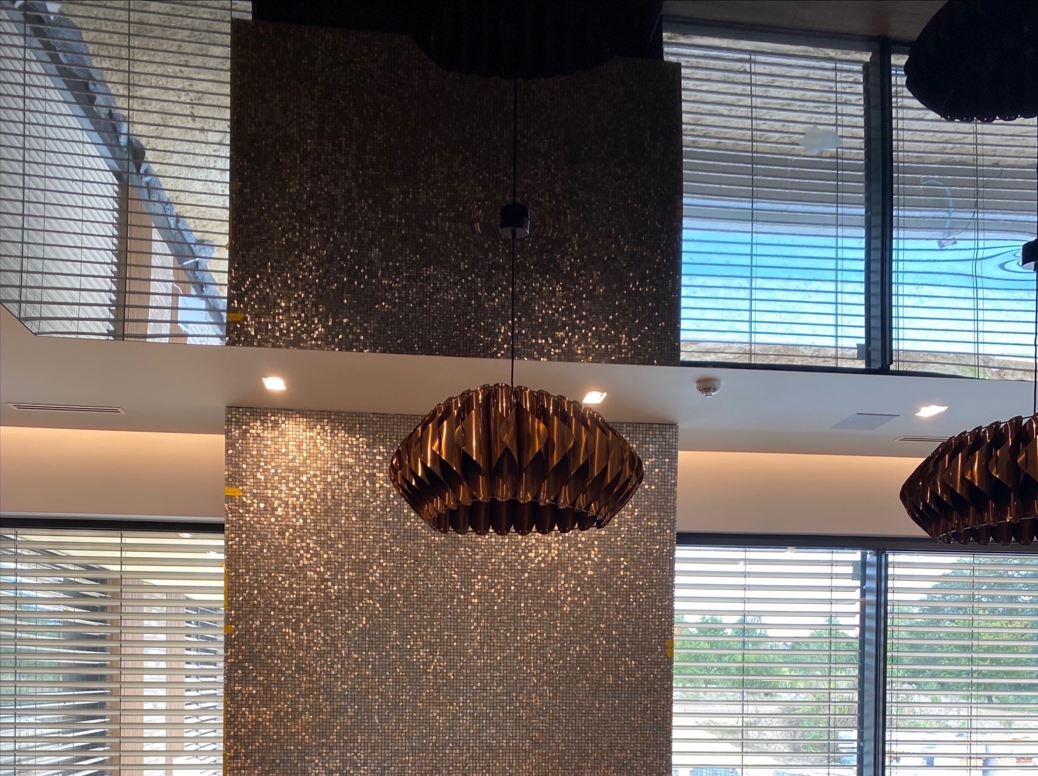 Mozaika z połyskujących płytek na ścianie, widok na okno z żaluzjami i dekoracyjna lampa wisząca z drewnianych elementów.