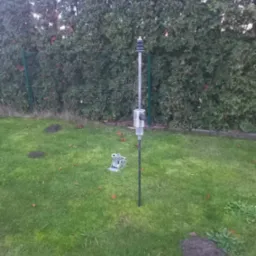 Instalacja do pomiaru gruntu na trawniku z kretowiskami, widoczne elementy metalowe i plastikowe, w tle żywopłot.