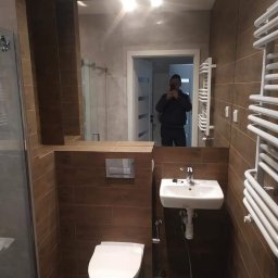 HIT HOUSE - Wykończona łazienka z brązowymi płytkami imitującymi drewno, wiszącą toaletą, umywalką i grzejnikiem drabinkowym; w lustrze odbicie osoby robiącej zdjęcie.