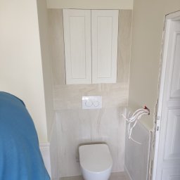 HIT HOUSE - Zamknięta toaleta podwieszana z szafką nad nią, białe płytki imitujące marmur na ścianie, widoczne kable elektryczne wystające ze ściany przed wykończeniem przy drzwiach.