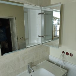 HIT HOUSE - Łazienka w trakcie remontu: zamontowana szafka z lustrem nad umywalką, widoczne wystające kable elektryczne i fragment glazury.