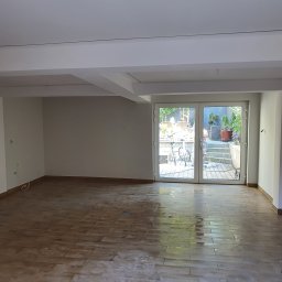 HIT HOUSE - Surowe wnętrze z drewnopodobną podłogą w trakcie czyszczenia, widoczne plamy wody i refleksy świetlne, duże przeszklone drzwi balkonowe z widokiem na ogród z rowerem.