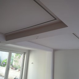 HIT HOUSE - Nowoczesne wnętrze: biały sufit z podwieszanymi elementami i listwami, widok na okno z zielenią, świeżo pomalowane ściany.