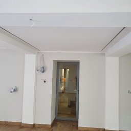 HIT HOUSE - Wnętrze w trakcie remontu: białe ściany, drewniana podłoga, szklane drzwi z szarą ramą, wystające kable elektryczne ze ściany i sufitu, listwy sufitowe.