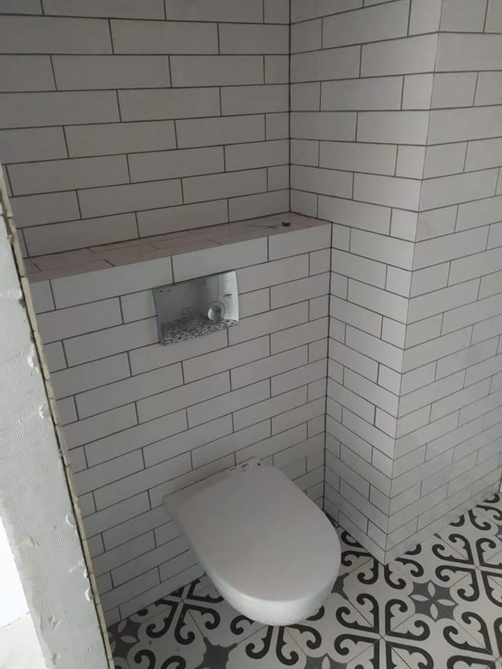 Świeżo wykafelkowana toaleta z białymi, prostokątnymi płytkami ułożonymi w cegiełkę, z widoczną spłuczką podtynkową i miską ustępową na podłodze z wzorzystych, szaro-czarnych płytek.