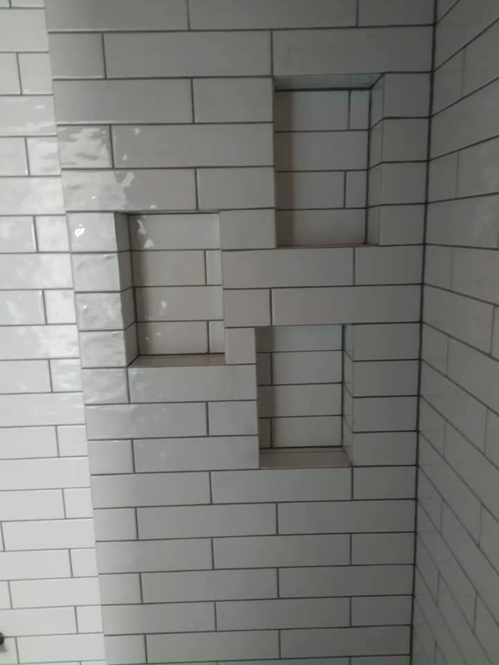 Ściana łazienki wyłożona białymi, prostokątnymi płytkami z wnękami na kosmetyki, ułożonymi w geometryczny wzór.