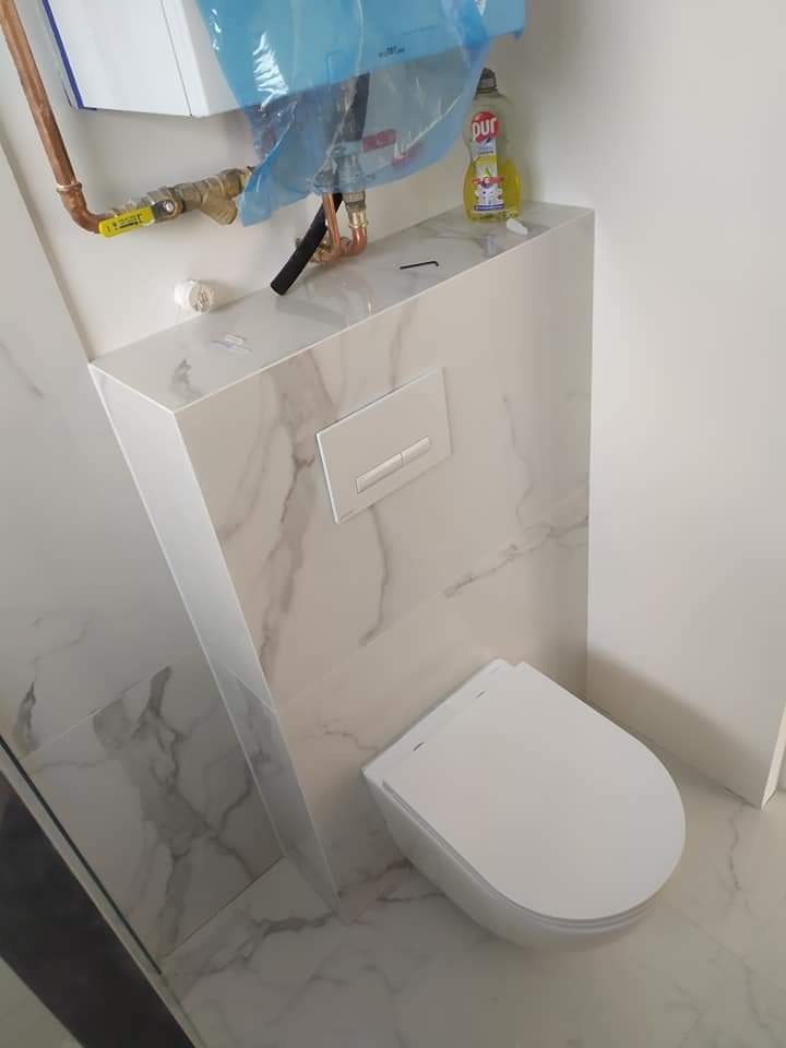 Nowoczesna toaleta podwieszana z marmurowymi płytkami, widoczna instalacja wodna i butelka płynu do naczyń na zabudowie.