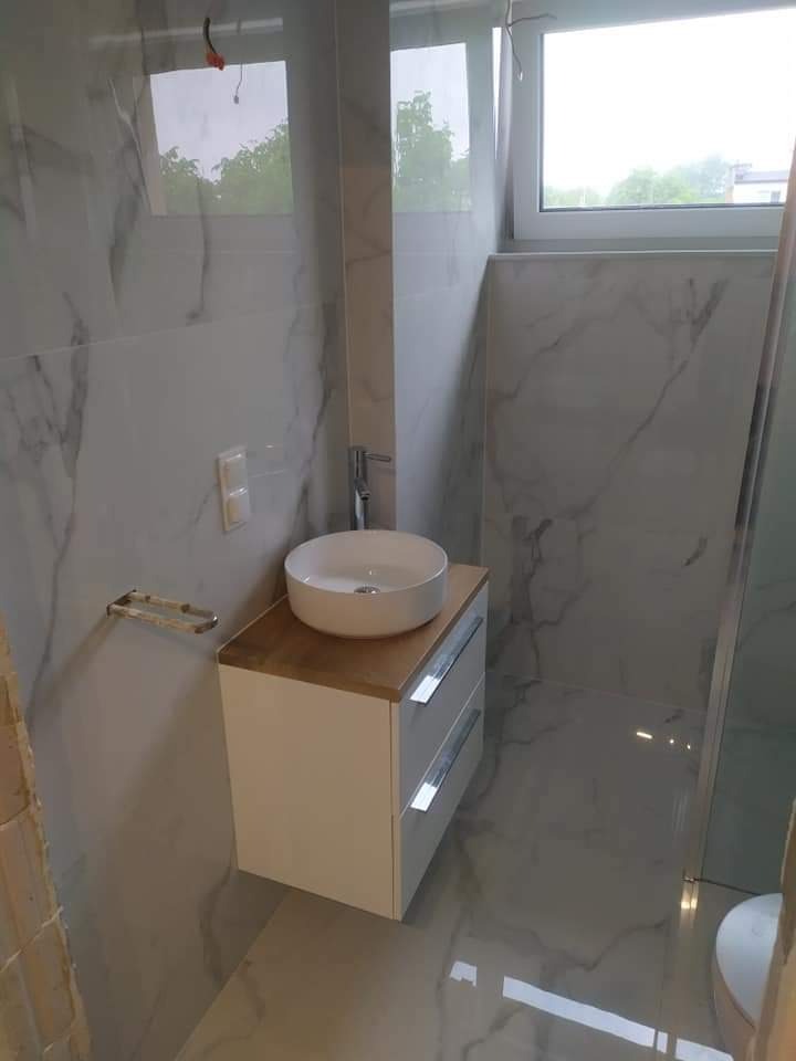 Nowoczesna łazienka z marmurowymi płytkami, okrągłą umywalką nablatową na białej szafce z drewnianym blatem i kabiną prysznicową ze szklaną ścianką.