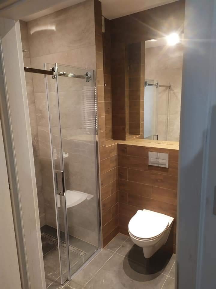Nowoczesna łazienka z kabiną prysznicową ze szklanymi drzwiami przesuwnymi, wiszącą toaletą i brązowymi płytkami imitującymi drewno na ścianie oraz szarymi płytkami na podłodze.