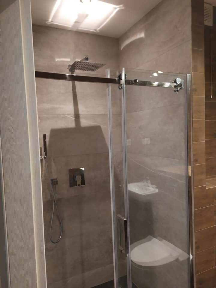 Nowoczesna kabina prysznicowa z deszczownicą i szklanymi drzwiami przesuwnymi, wykończona płytkami w odcieniach szarości i beżu, z widocznym fragmentem toalety wewnątrz kabiny.