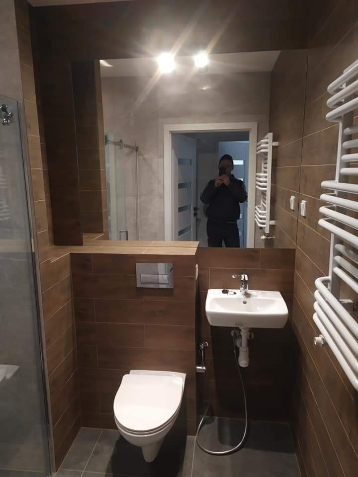 Wykończona łazienka z brązowymi płytkami imitującymi drewno, wiszącą toaletą, umywalką i grzejnikiem drabinkowym; w lustrze odbicie osoby robiącej zdjęcie.