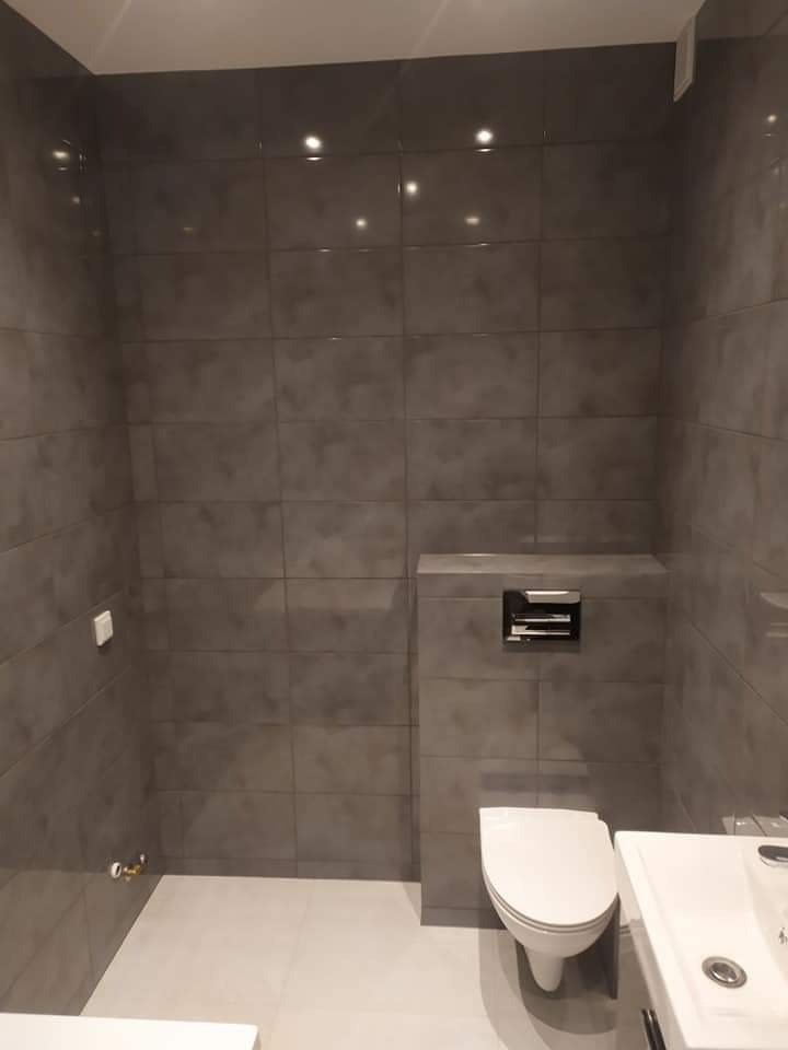 Nowoczesna łazienka z szarymi, matowymi płytkami wielkoformatowymi, podwieszanym WC i minimalistyczną umywalką, oświetlona punktowymi lampami sufitowymi.