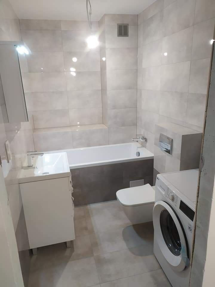 Jasna łazienka z wanną w zabudowie, umywalką z szafką, toaletą i pralką, wykończona płytkami w odcieniach szarości, z widoczną kratką wentylacyjną pod sufitem.