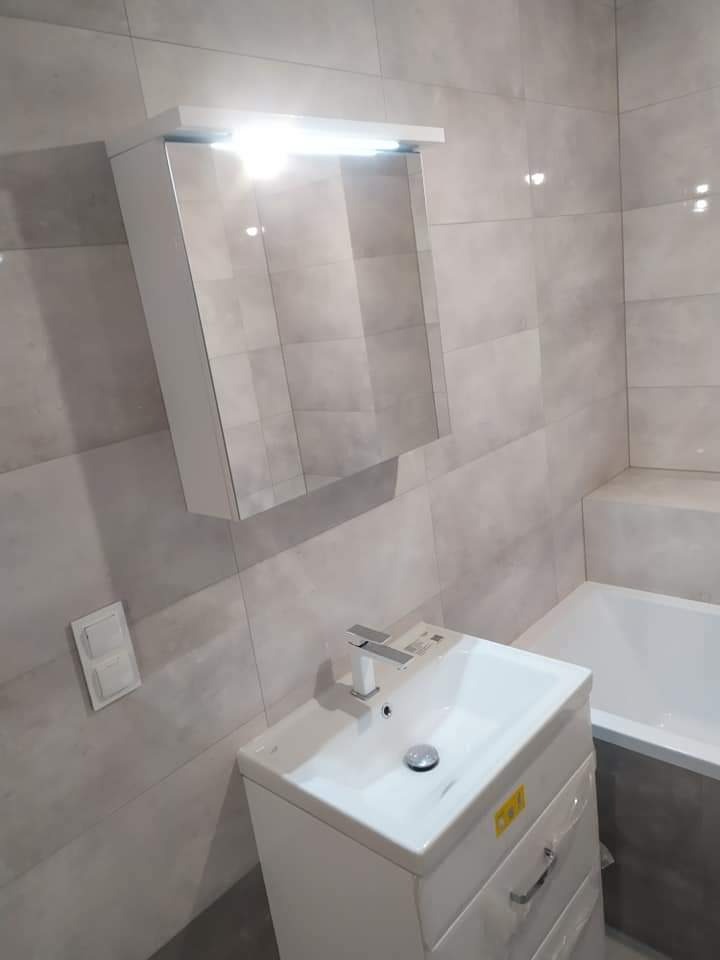 Nowoczesna łazienka z jasnymi, kwadratowymi płytkami, białą szafką z umywalką i lustrem z oświetleniem LED, minimalistyczna bateria, widoczny fragment wanny.