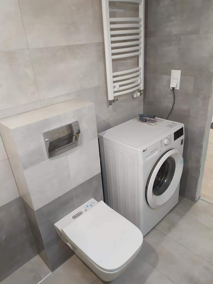 Nowoczesna łazienka z szarymi płytkami, podwieszaną toaletą z funkcją bidetu, grzejnikiem drabinkowym i pralką LG.