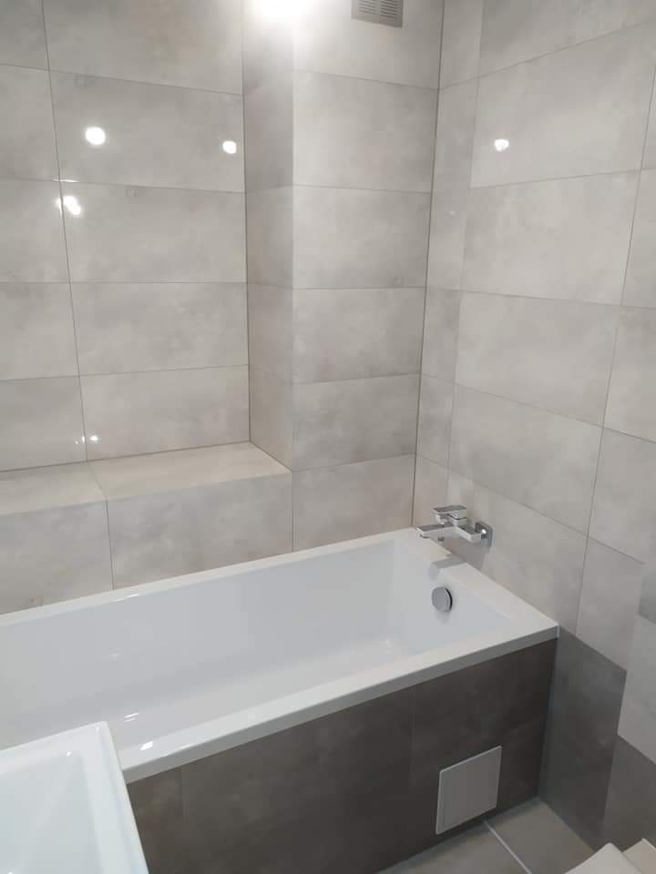 Nowoczesna łazienka z wanną obudowaną szarymi płytkami, jasnoszare płytki na ścianach, chromowana bateria, widoczny fragment umywalki.