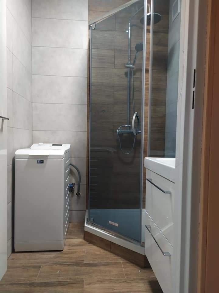 Nowoczesna łazienka z kabiną prysznicową narożną z przezroczystego szkła, białą pralką i umywalką z szafką w odcieniach bieli i drewna, wykończona płytkami w kolorach szarości i brązu imitującymi...