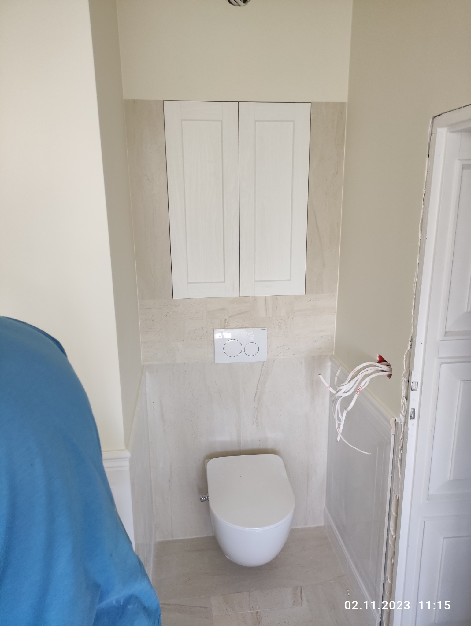 Zamknięta toaleta podwieszana z szafką nad nią, białe płytki imitujące marmur na ścianie, widoczne kable elektryczne wystające ze ściany przed wykończeniem przy drzwiach.