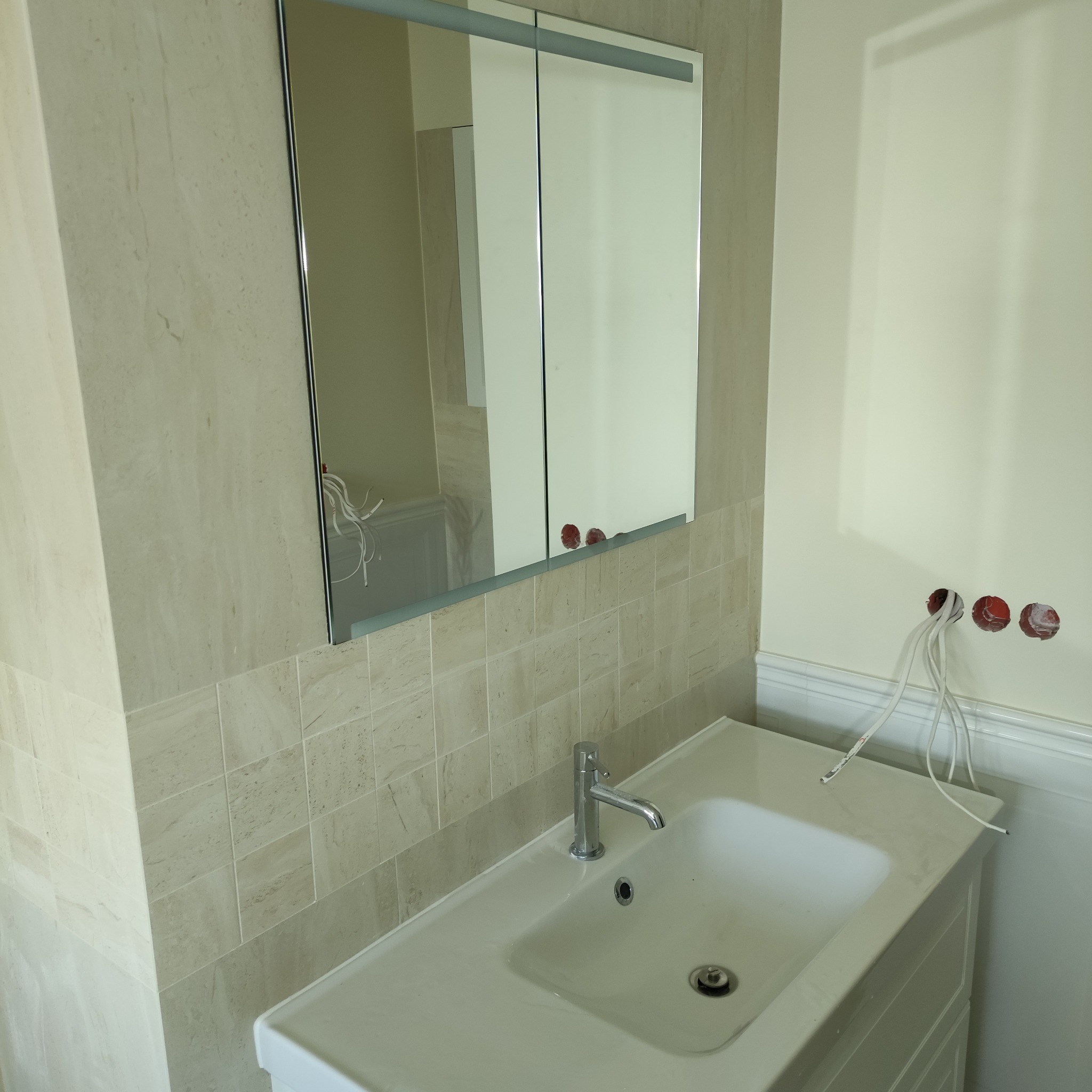 Umywalka z szafką w łazience wyłożonej beżowymi płytkami, widoczne niezakończone podłączenia elektryczne i lustro z szafką.