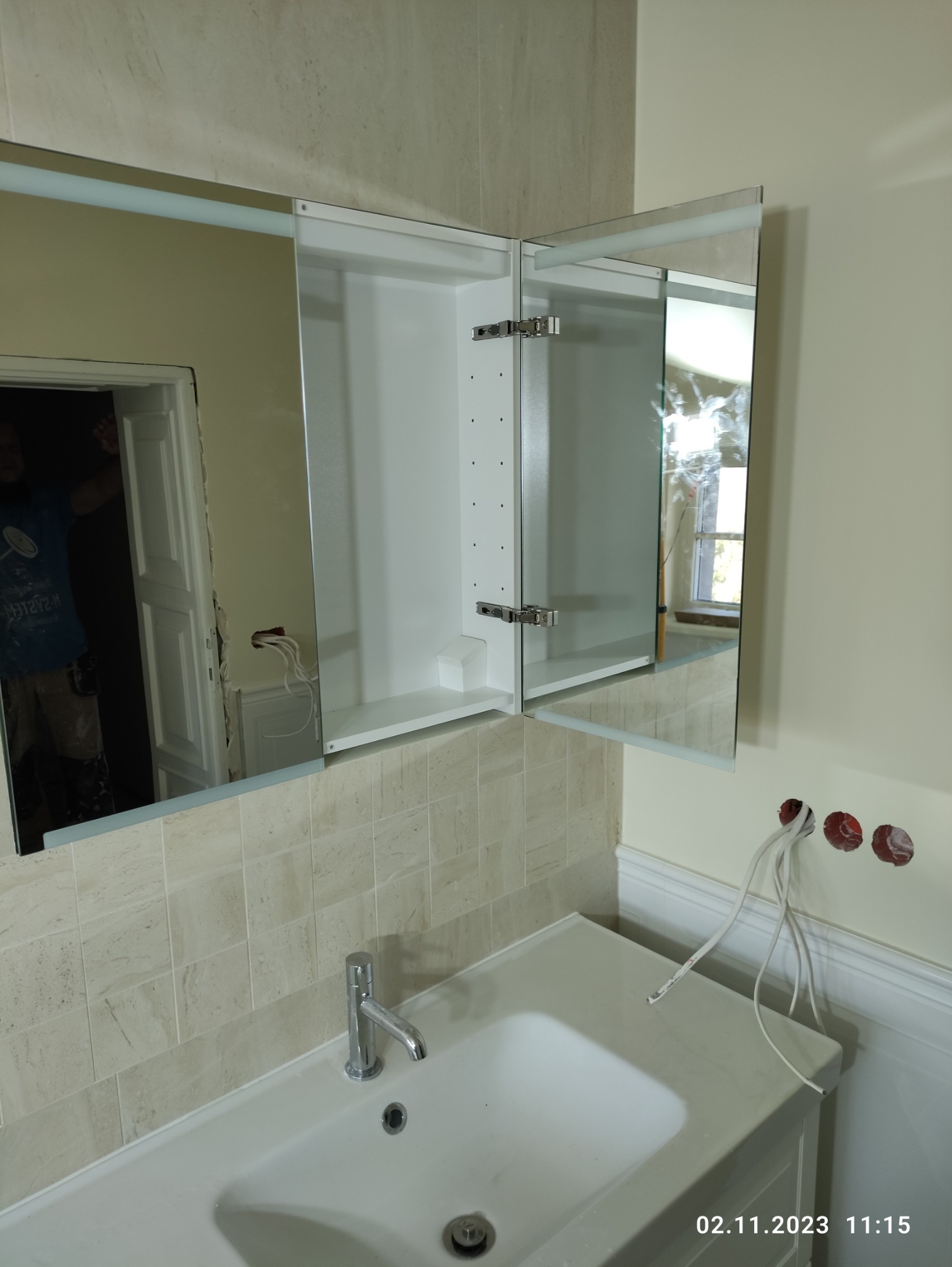 Łazienka w trakcie remontu: zamontowana szafka z lustrem nad umywalką, widoczne wystające kable elektryczne i fragment glazury.