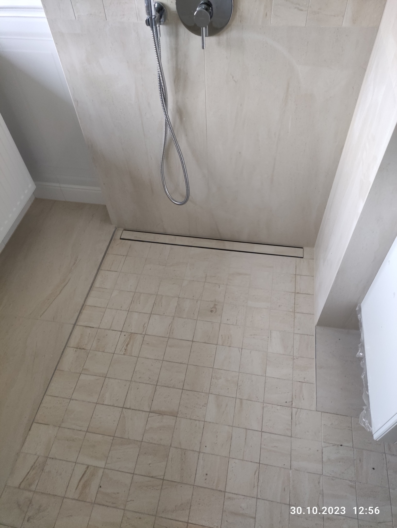 Wykończona kabina prysznicowa z beżowymi płytkami, odpływ liniowy w posadzce, bateria natryskowa zamontowana na ścianie.