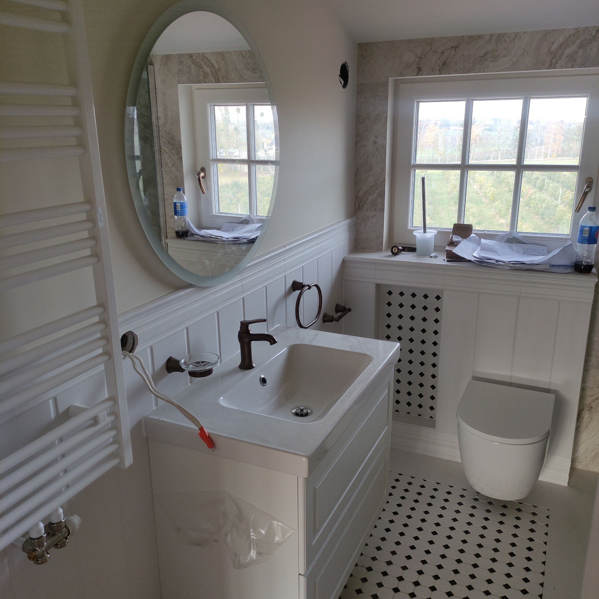 Biała łazienka w trakcie wykańczania: umywalka z brązową armaturą, grzejnik drabinkowy z wystającym kablem, okrągłe lustro, toaleta i czarno-biała podłoga w romby. Widoczne narzędzia i materiały...