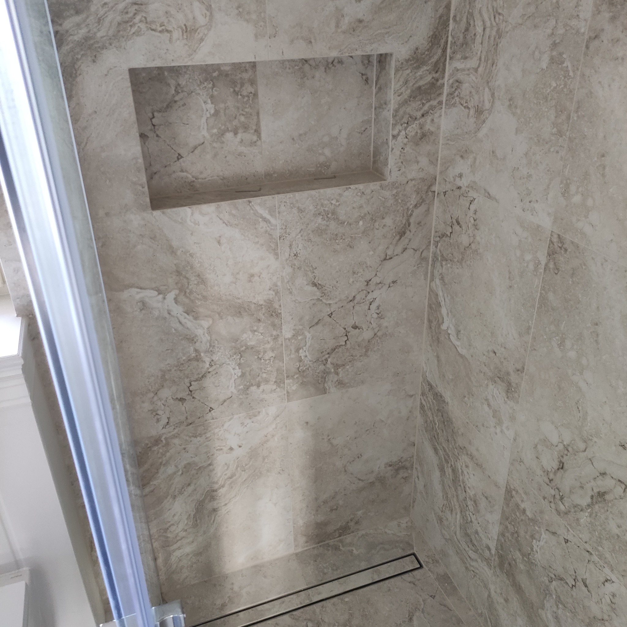 Wykończona kabina prysznicowa z szarymi płytkami imitującymi marmur i wnęką na kosmetyki, widoczny odpływ liniowy w podłodze i fragment szklanej ściany.