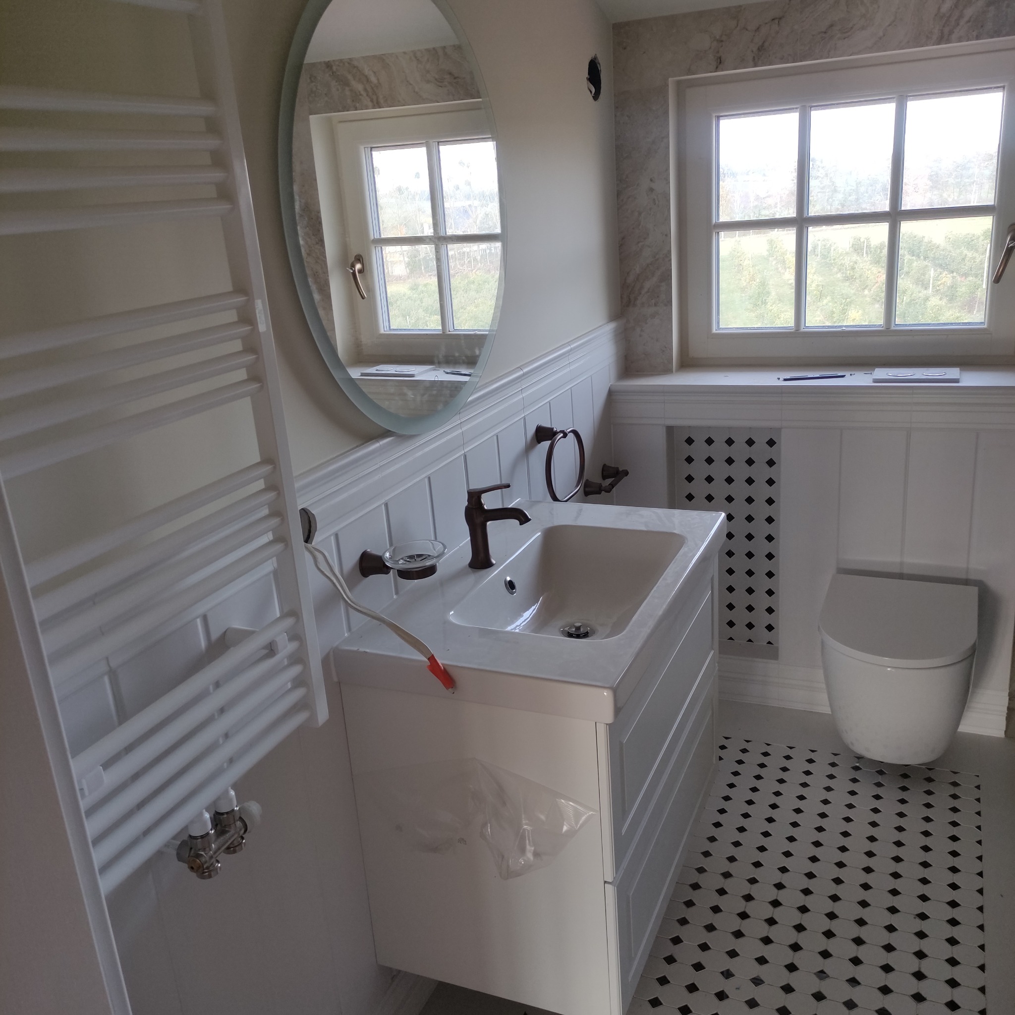 Wykończona łazienka w stylu klasycznym z białą armaturą, grzejnikiem drabinkowym, umywalką z szafką, toaletą i podłogą w czarno-białą szachownicę. Widoczne okrągłe lustro i okno z widokiem...