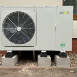 Biała pompa ciepła Nova, model R32, zamontowana na betonowych blokach przed budynkiem z ceglaną podmurówką i brązowym oknem. Widoczne rury i kamienie.