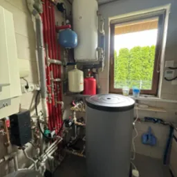 Złożona instalacja grzewcza z szarym zasobnikiem ciepłej wody, czerwonymi rurami, białym bojlerem i niebieskim naczyniem wzbiorczym przy oknie z widokiem na zieleń.