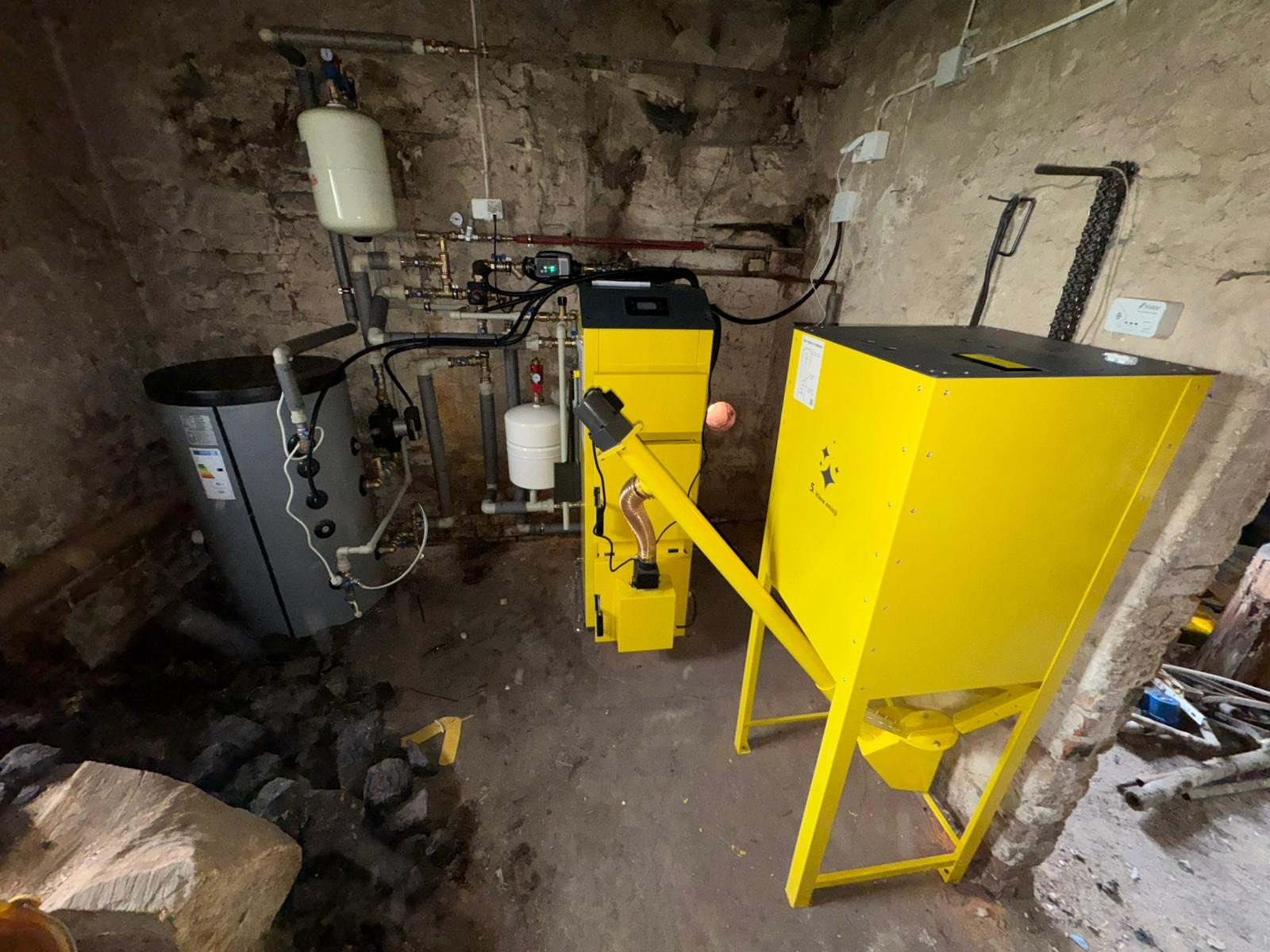 Żółty piec na paliwo stałe z podajnikiem ślimakowym w piwnicy o kamiennych ścianach, obok szary zasobnik wody. Widoczne rury i instalacje.