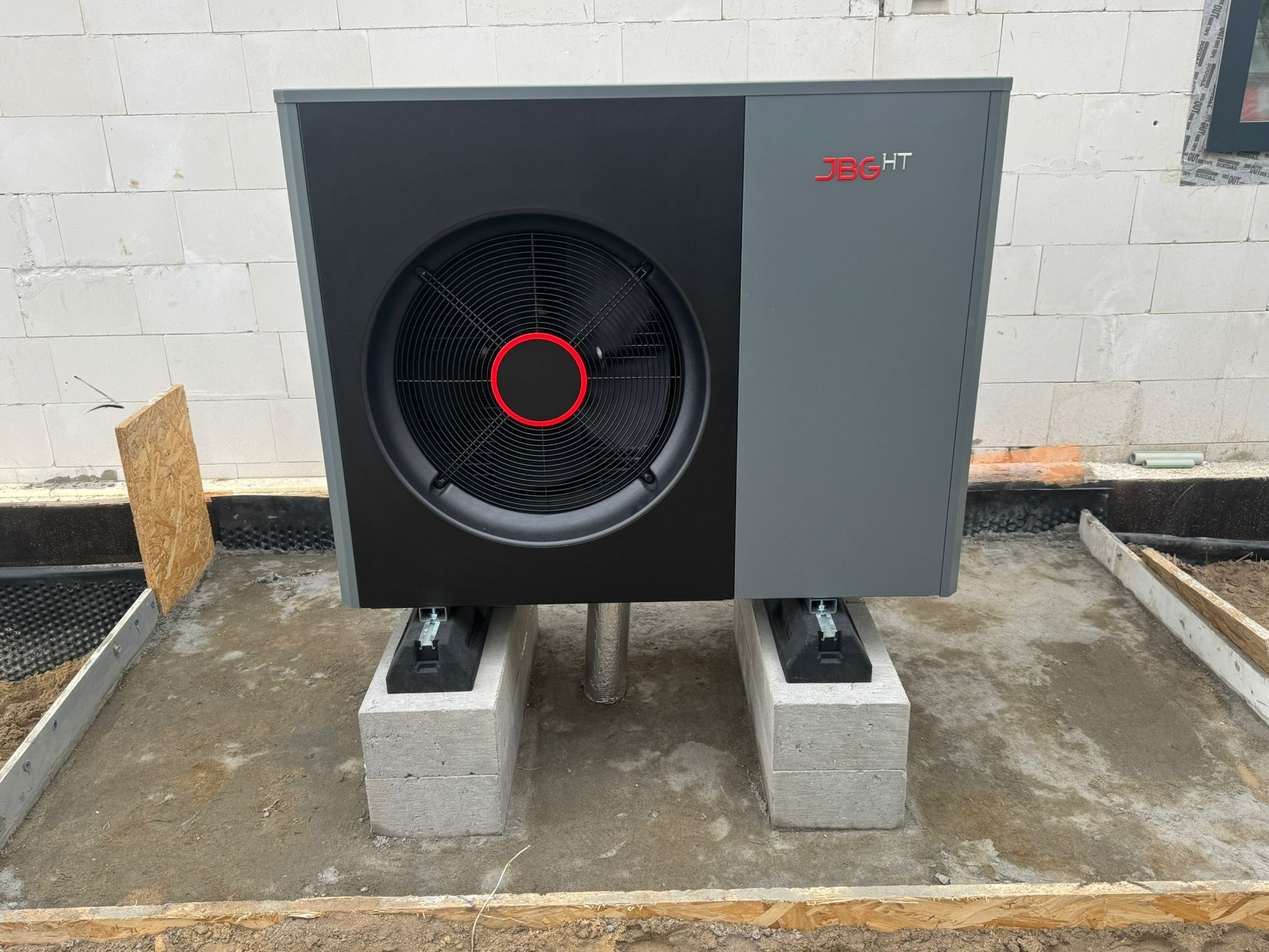Szara pompa ciepła JBG HT na betonowych blokach fundamentowych, widok z przodu na tle ściany z białych bloczków. Montaż na przygotowanym podłożu.