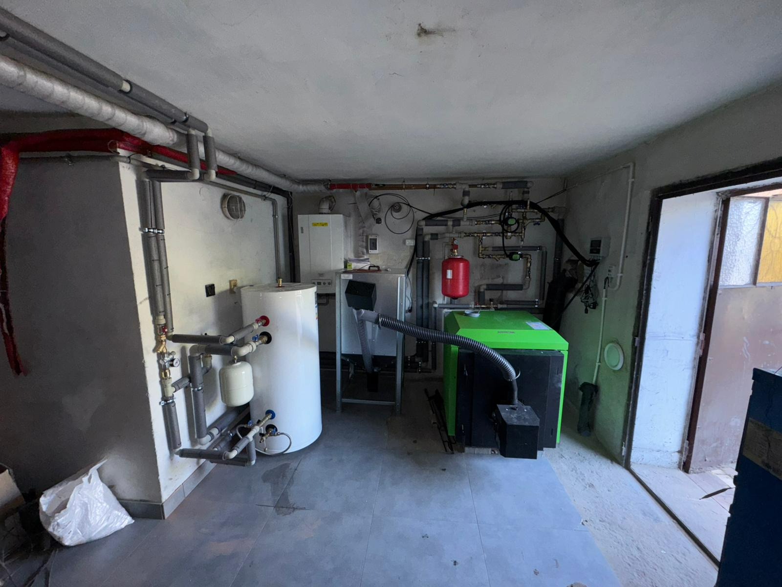 Kotłownia z zielonym piecem na paliwo stałe, białym zbiornikiem i rozbudowaną instalacją hydrauliczną. Widoczne rury, zawory i inne elementy. Szare płytki na podłodze.