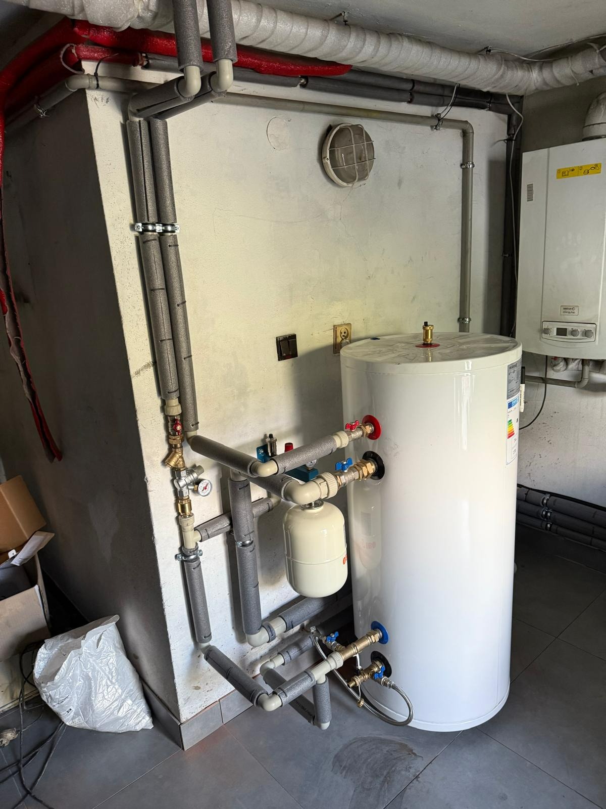 Biały, cylindryczny zbiornik ciepłej wody użytkowej z podłączeniami hydraulicznymi, widoczne rury w izolacji, manometr. Na ścianie piec gazowy.