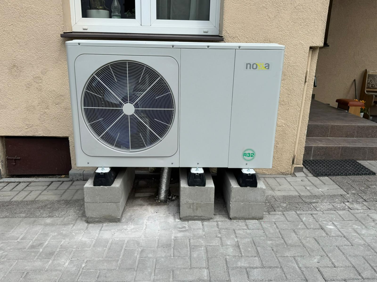 Zamontowana pompa ciepła marki Nova, ustawiona na betonowych bloczkach przy ścianie budynku. Widoczne okno i wejście do domu. Oznaczenie R32.