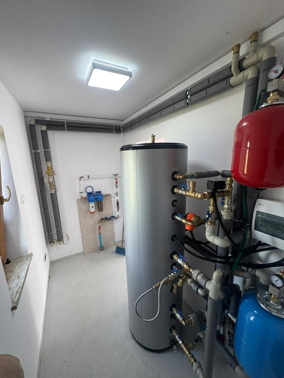 Szara, pionowa instalacja grzewcza z licznymi zaworami i rurami w schludnym pomieszczeniu z szarymi rurami przy suficie i filtrami wody na ścianie.