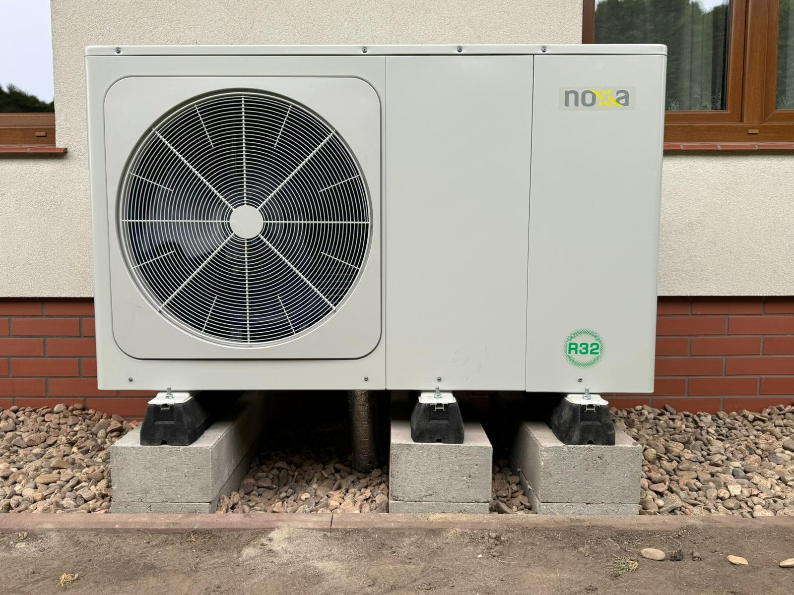 Biała pompa ciepła Nova, model R32, zamontowana na betonowych blokach przed budynkiem z ceglaną podmurówką i brązowym oknem. Widoczne rury i kamienie.