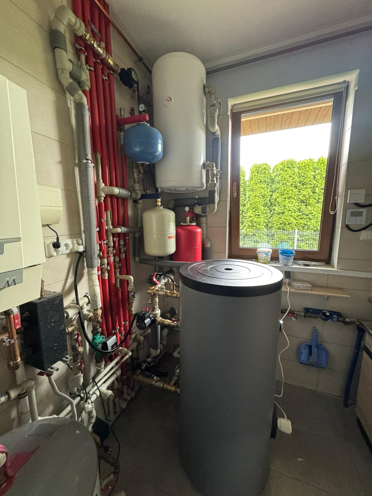 Złożona instalacja grzewcza z szarym zasobnikiem ciepłej wody, czerwonymi rurami, białym bojlerem i niebieskim naczyniem wzbiorczym przy oknie z widokiem na zieleń.