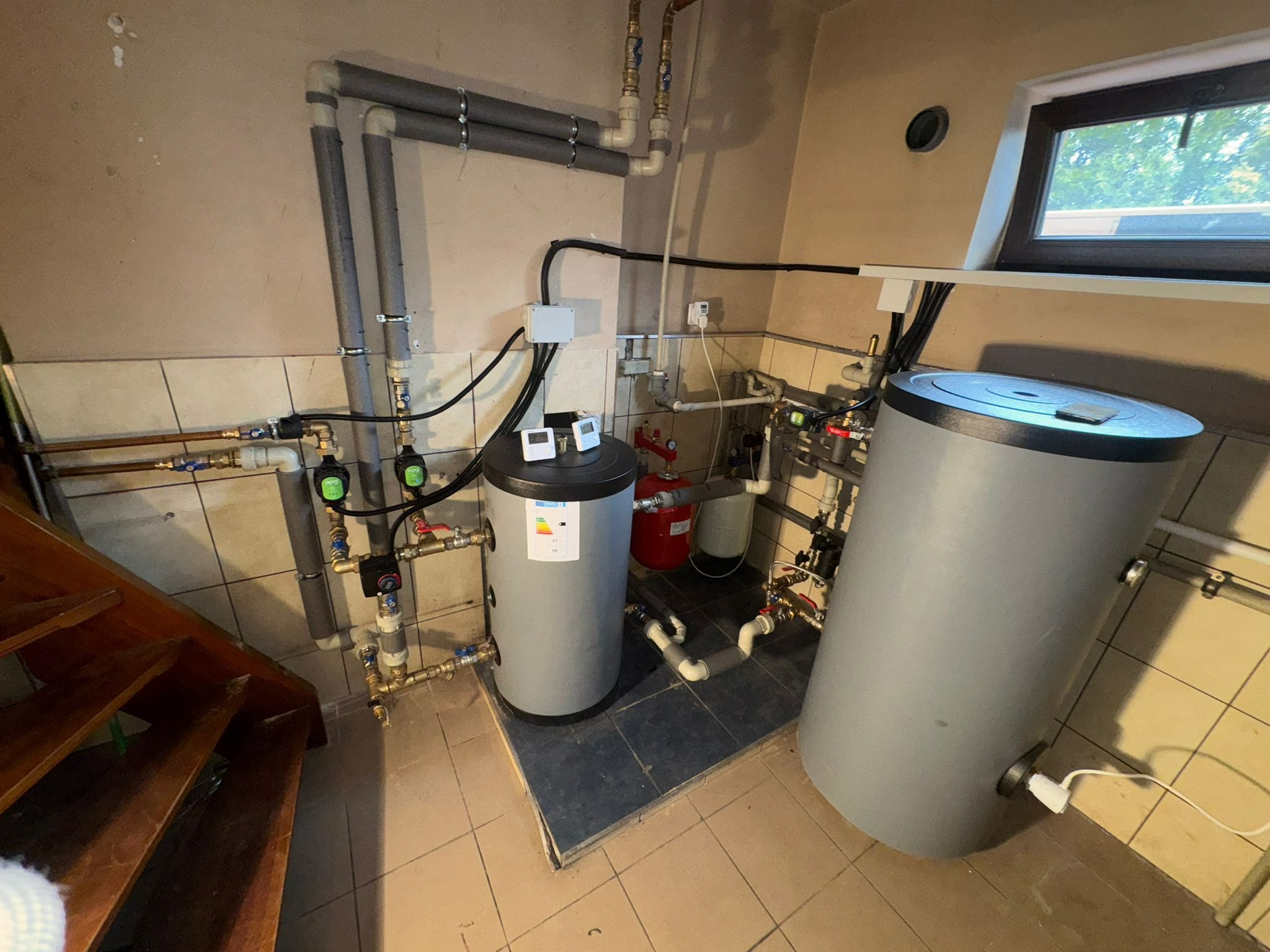 Dwa szare zasobniki ciepłej wody użytkowej w kotłowni, z widocznymi rurami, zaworami i elementami instalacji grzewczej. Ujęcie z perspektywy schodów.