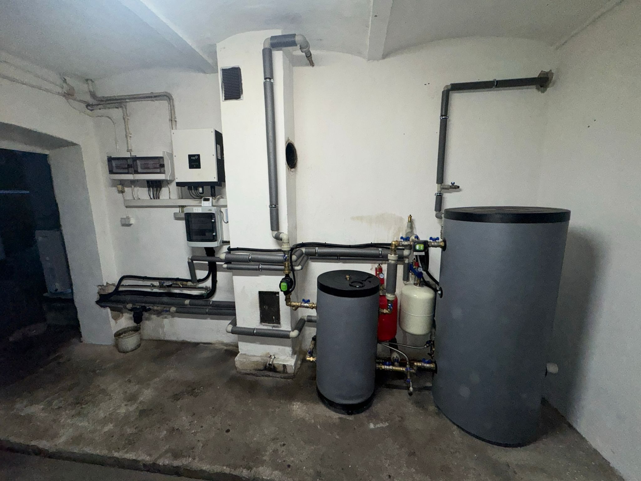 Złożona instalacja grzewcza w piwnicy: dwa szare zbiorniki na wodę, biała pompa ciepła, rury z izolacją. Widoczne liczniki i okablowanie. Surowy beton na podłodze.