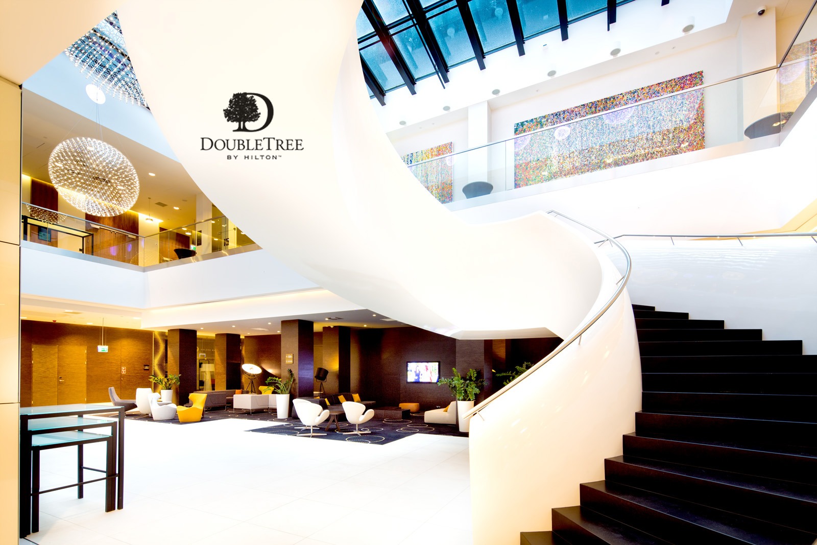Przestronne lobby hotelowe z białą spiralną klatką schodową, nowoczesnymi lampami wiszącymi i komfortową strefą wypoczynkową z sofami i fotelami.