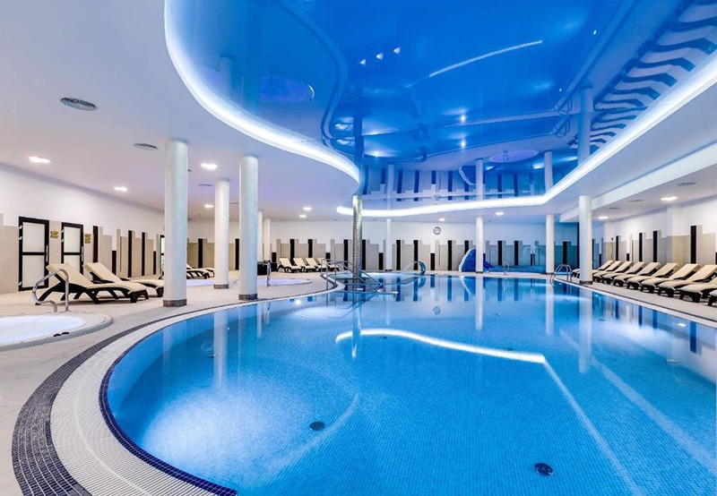 Błękitny sufit napinany z oświetleniem LED nad basenem w nowoczesnym spa, odbicie w tafli wody, białe kolumny i leżaki w tle.